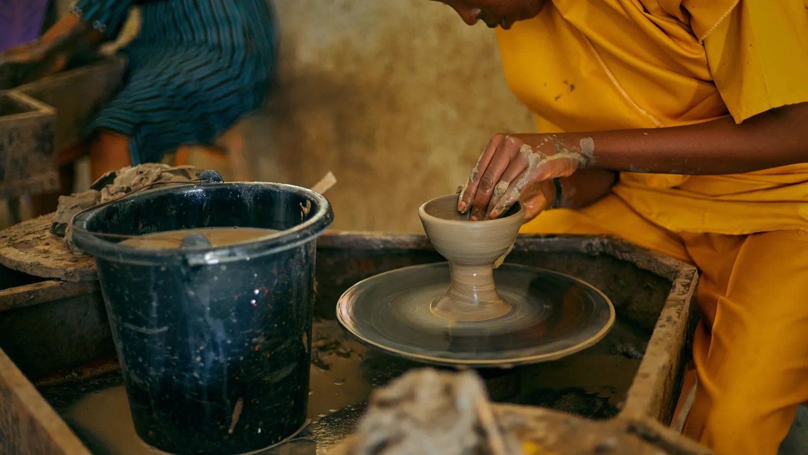 Photo: Finalchoice on Pexels A potter in a yellow smock shaping a small clay cup on a wheel, in a busy workshop